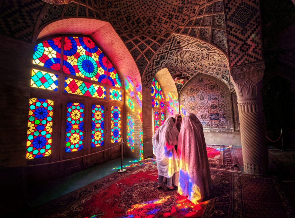 Women in traditional attire inside the vibrant Nasir al-Mulk Mosque in Shiraz, Iran.